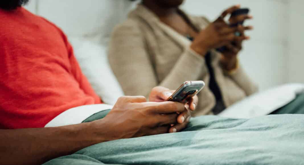 A close up of a man's hands as he looks at his phone in bed, a women is beside him out of focus.
